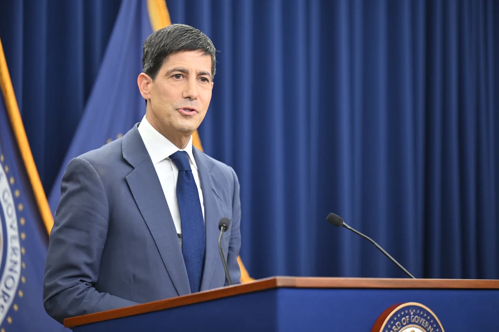 Chairman Warsh speaking at a podium with microphones in front of a blue backdrop.