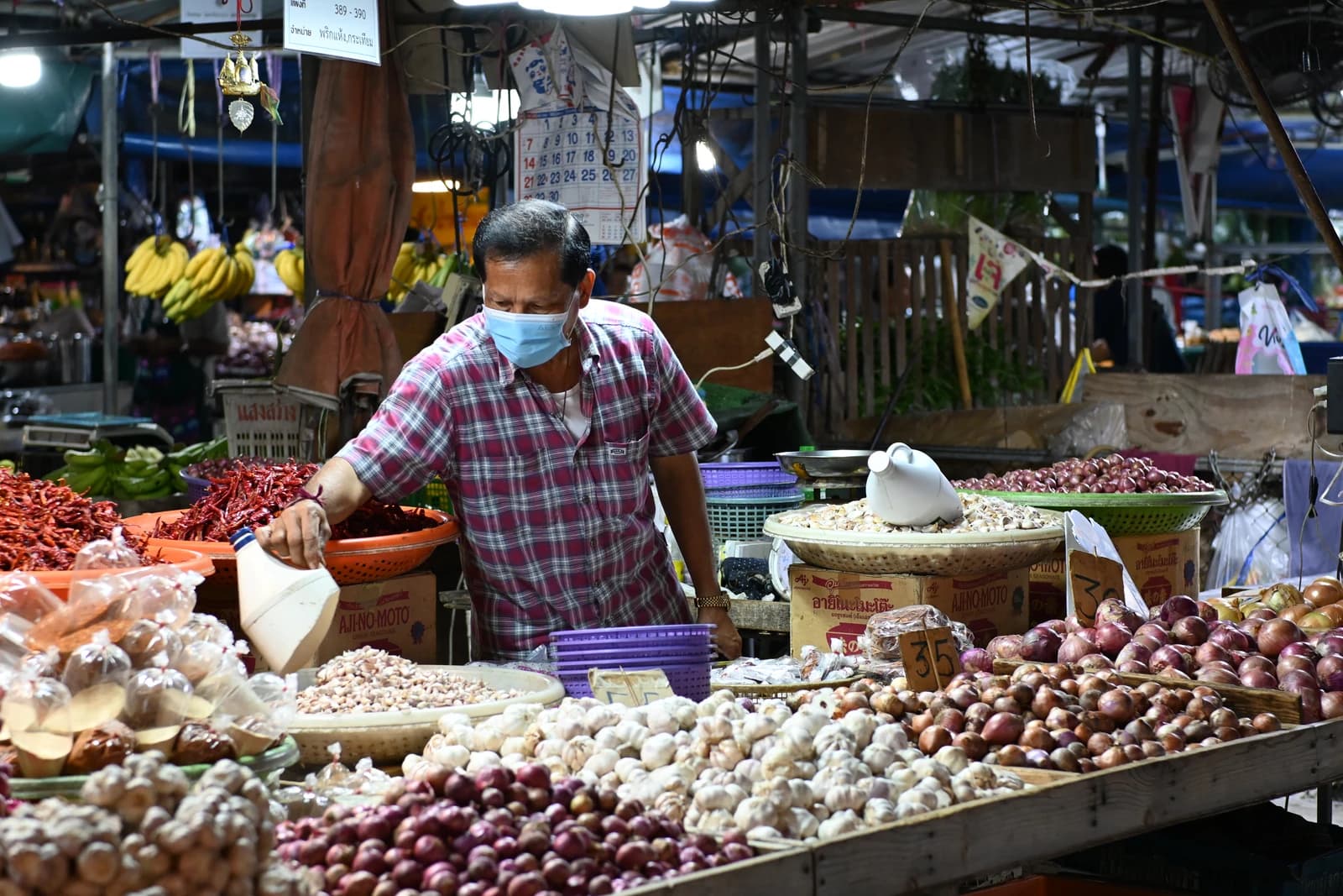 Masked vendor arranging baskets of garlic, shallots, and chilies at an outdoor market stall.