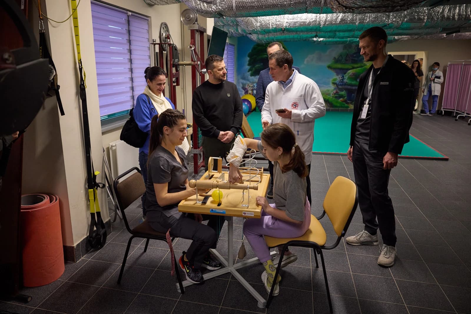 President Zelenskyy observing a child and therapist engaging with a rehabilitation device.