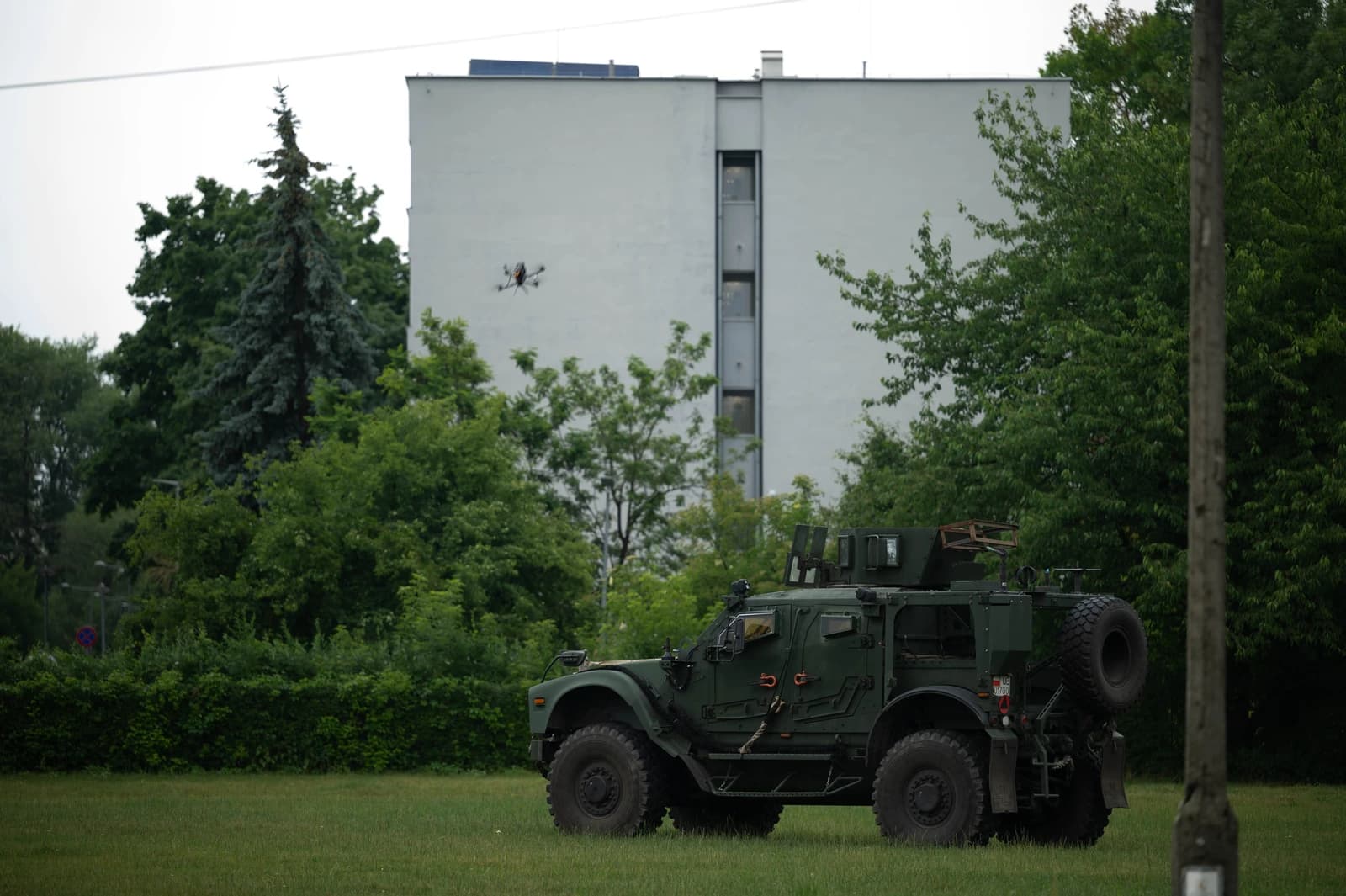 Military vehicle on grass with a drone in the air near a building.