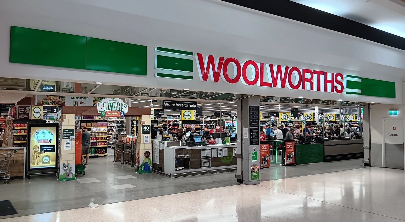 Entrance to a Woolworths supermarket with customers at self-checkout registers inside.