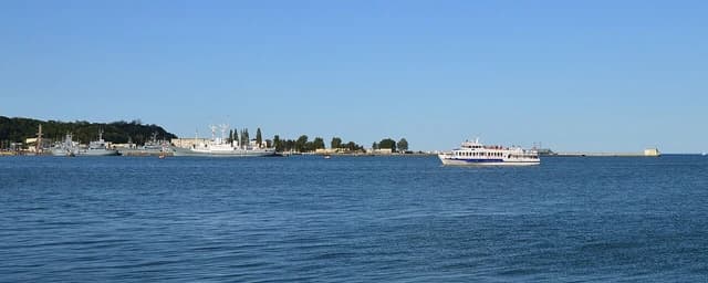 Several ships docked in a harbor with a passenger vessel sailing in the foreground.