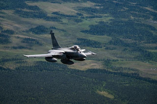 A grey Dassault Rafale fighter jet in flight over green forests and hills.