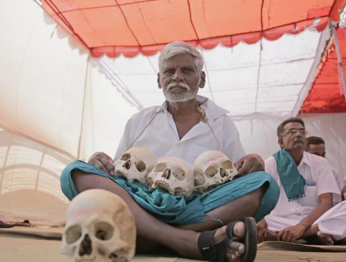 A farmer sits cross-legged holding three skulls at a protest.