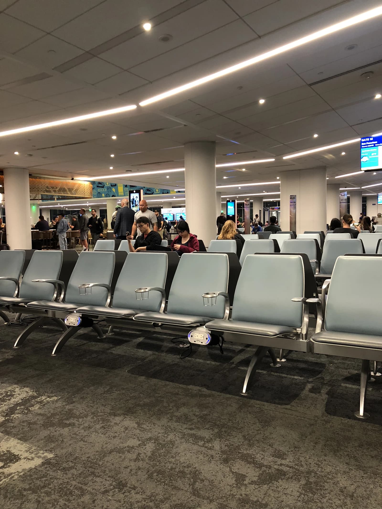 Travelers sit in waiting area chairs at a busy airport terminal.