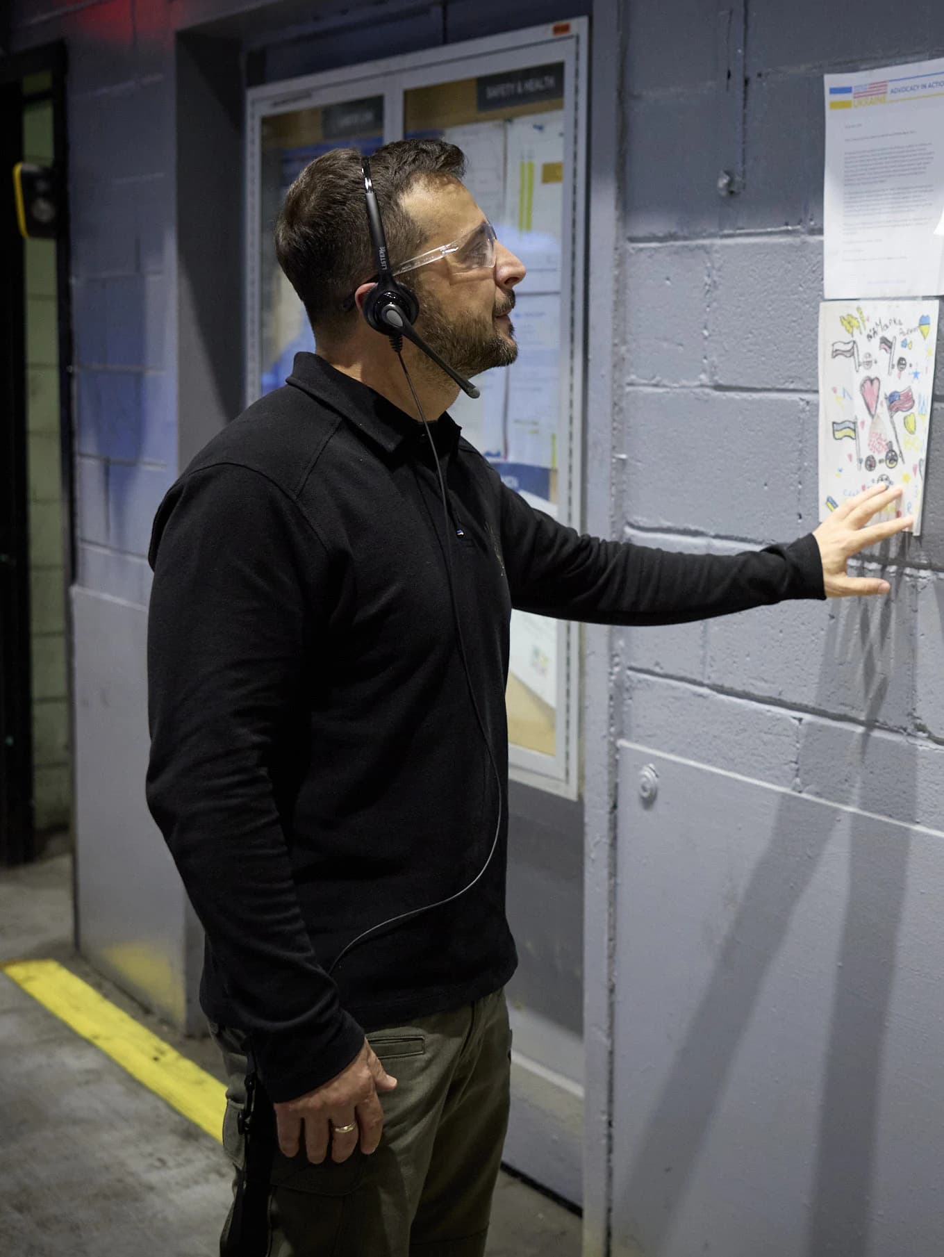 Volodymyr Zelenskyy, President of Ukraine, wearing safety glasses and a headset, touching a drawing.
