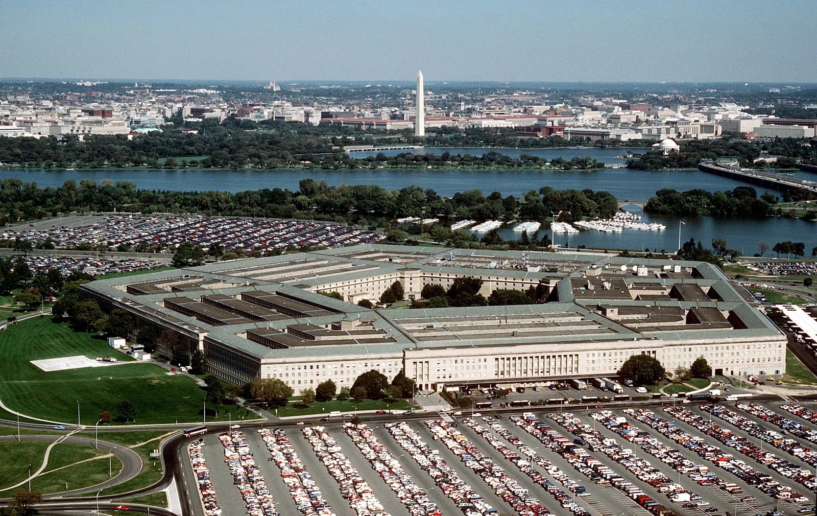 Aerial view of The Pentagon building with large parking lots, a river, and the Washington Monument in the distance.