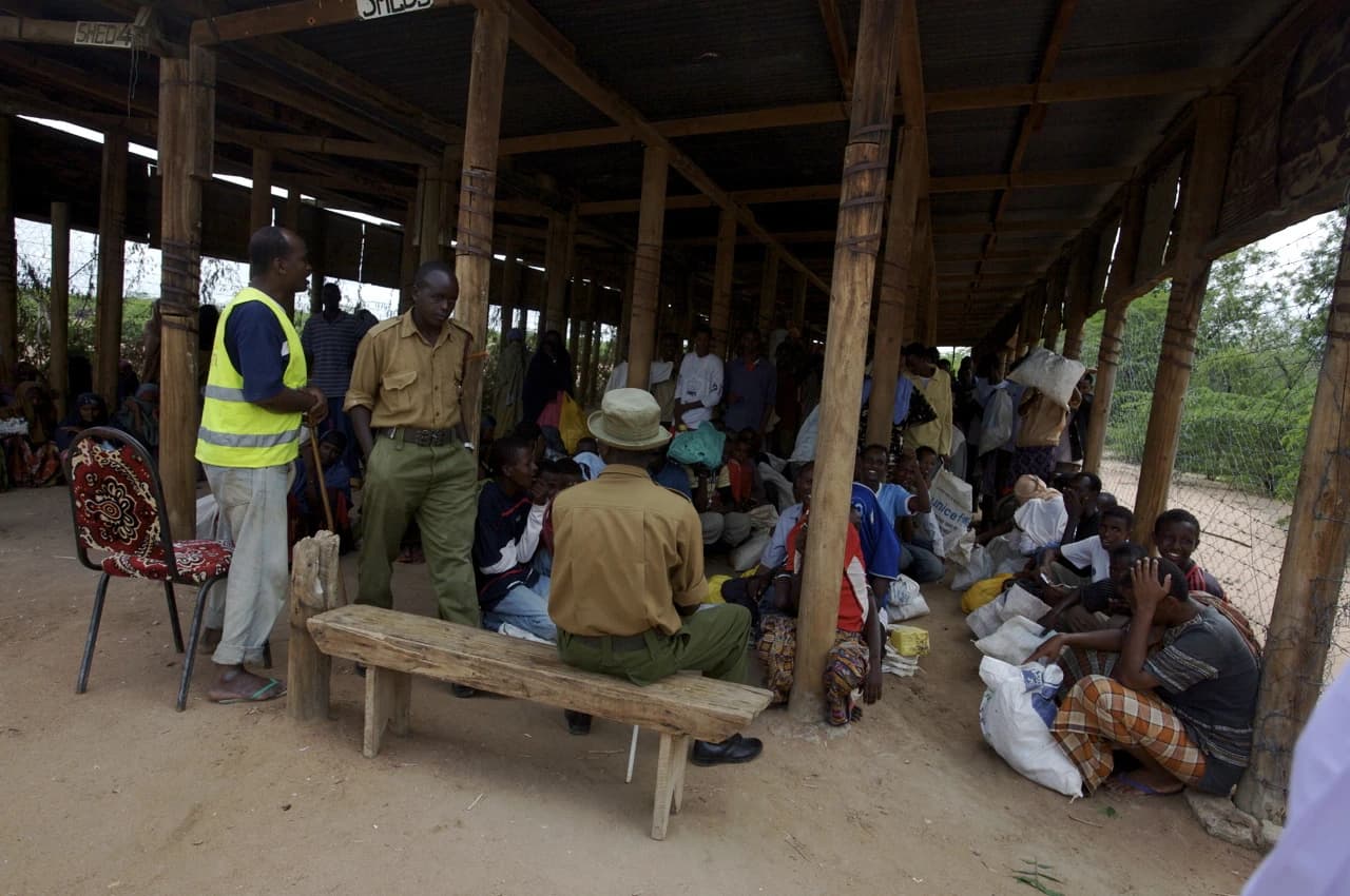 Group of people gathered under a wooden shelter, with aid workers and military personnel.