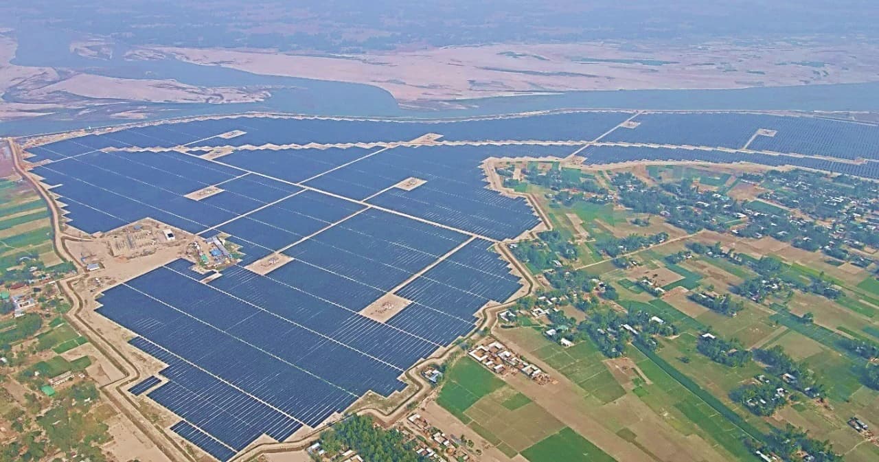 Aerial view shows a vast solar power plant with thousands of panels beside a wide river.