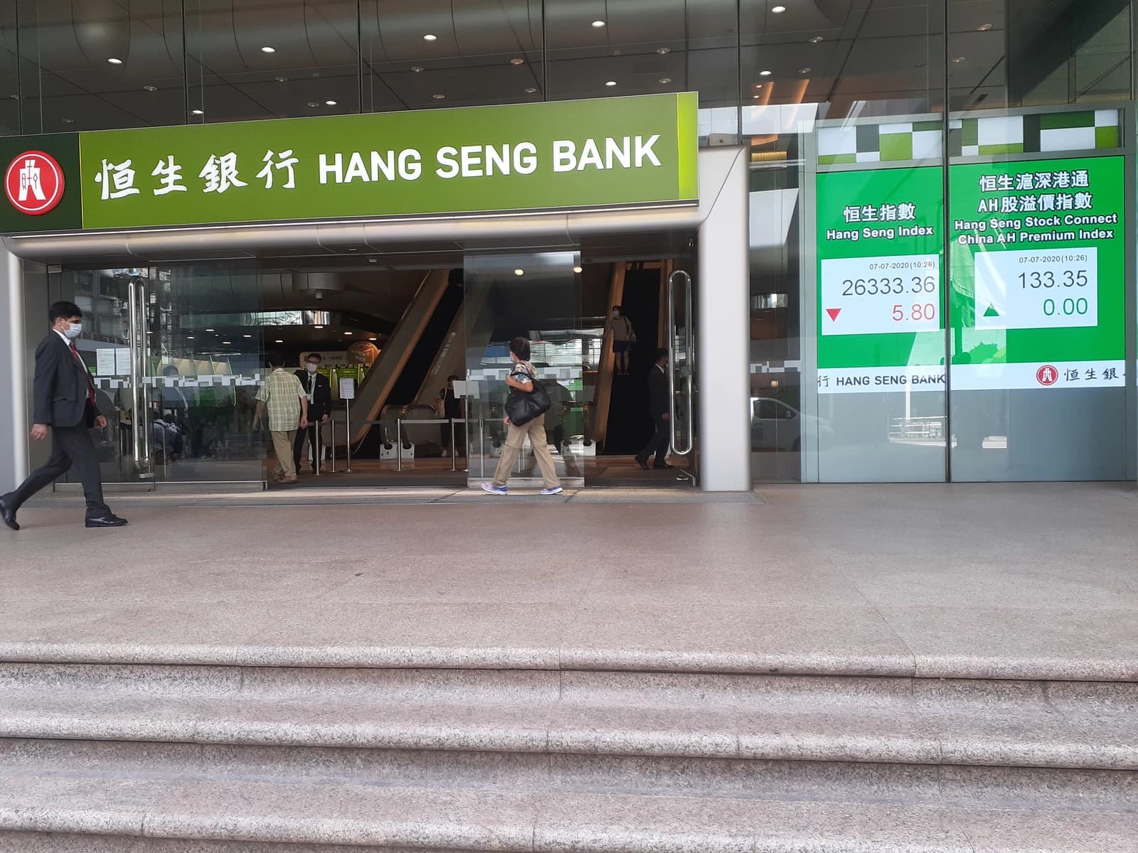 Pedestrians walk past the entrance of Hang Seng Bank with digital stock index displays.