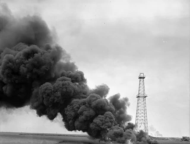An oil well fire emitting large plumes of dark smoke next to an oil derrick.
