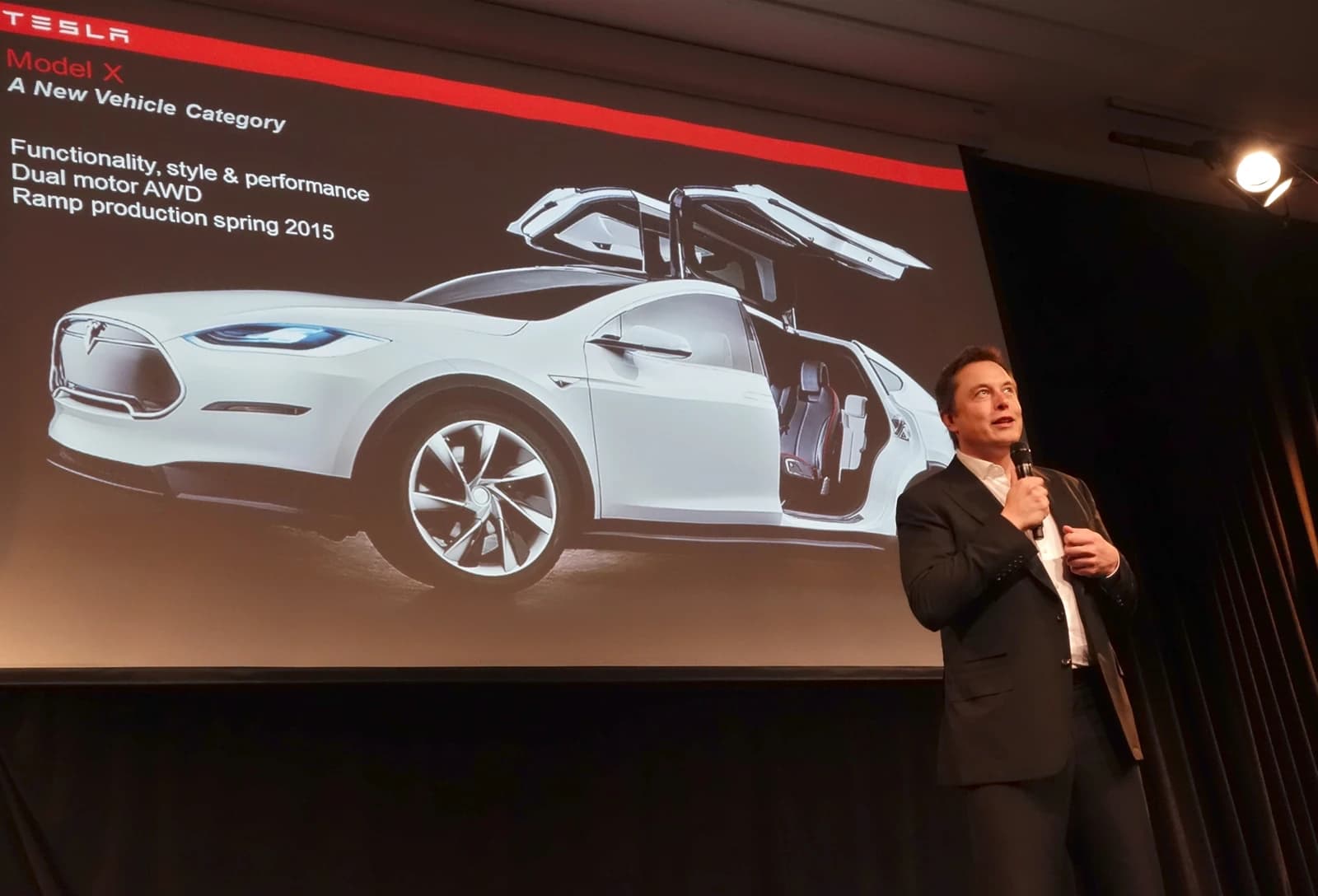 Elon Musk, CEO of Tesla, speaks on stage with a microphone in front of a screen displaying a Tesla Model X.