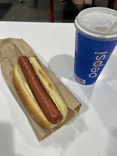 A hot dog in a bun next to a blue Pepsi cup on a white table.