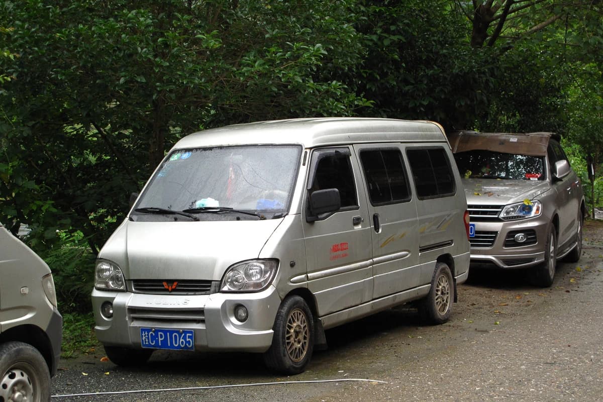 A silver Wuling Sunshine minivan parked on a wet, unpaved road in a wooded area.
