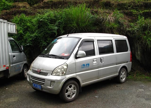 Silver Wuling Rongguang minivan parked on a gravel roadside next to dense foliage.