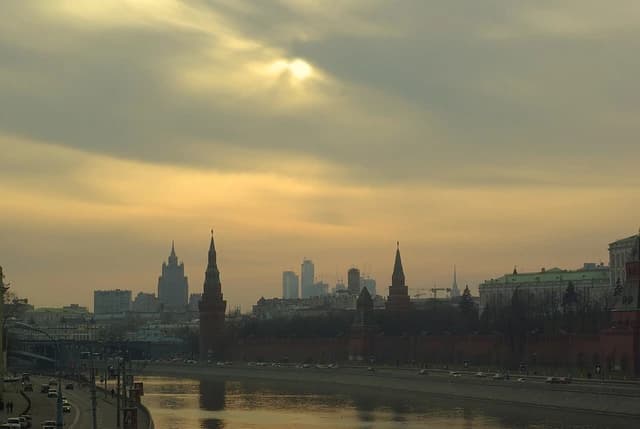 Cityscape of Moscow featuring the Kremlin towers, river, and skyline under a cloudy sky.