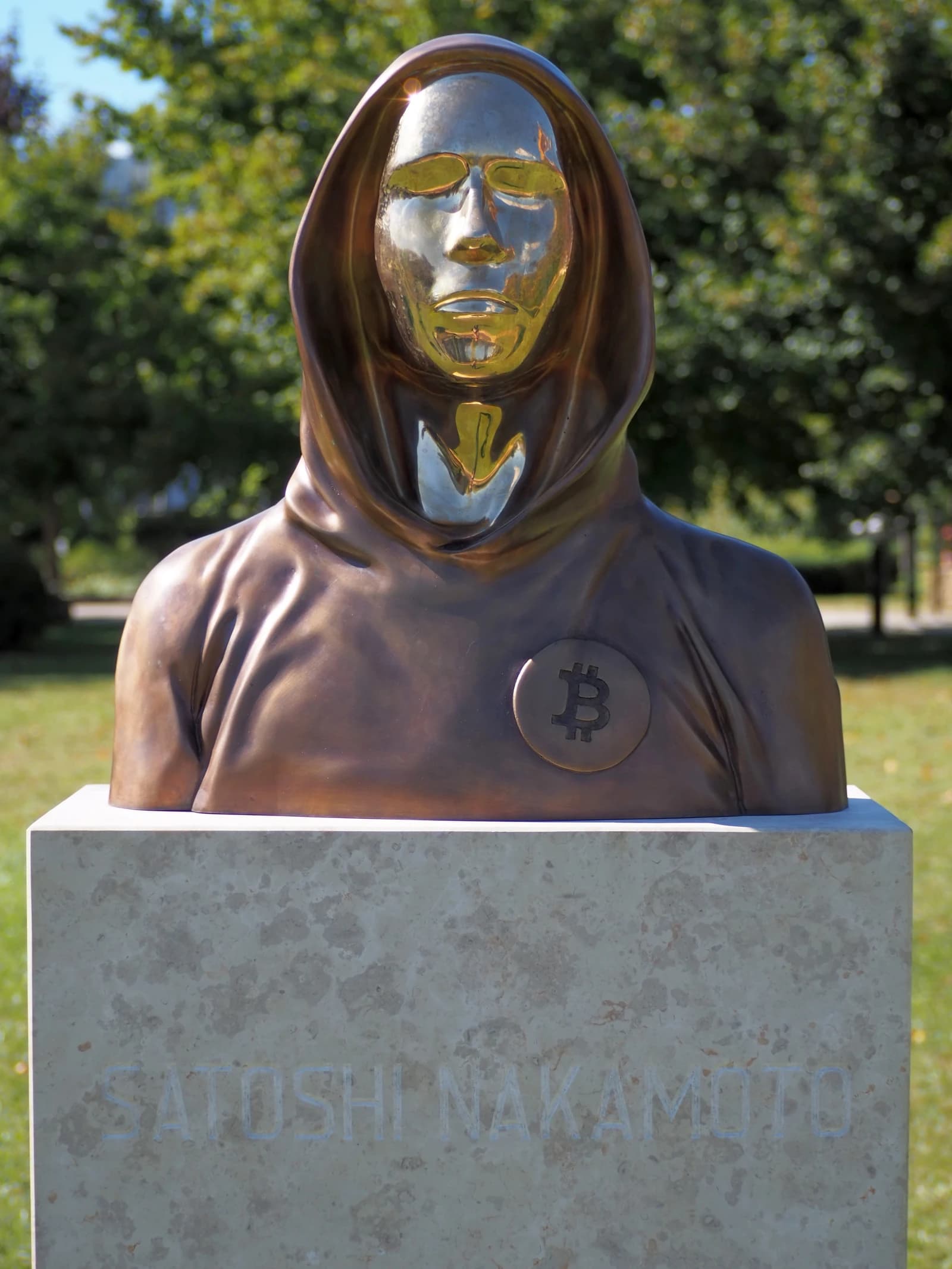Bust of Satoshi Nakamoto, creator of Bitcoin, in a park setting.