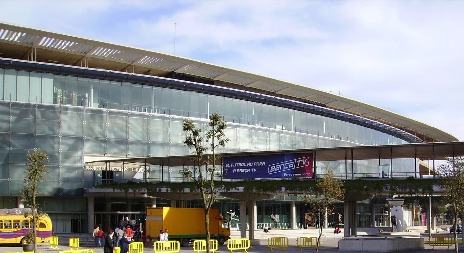 Exterior of the Camp Nou stadium with a yellow bus parked outside and people walking.