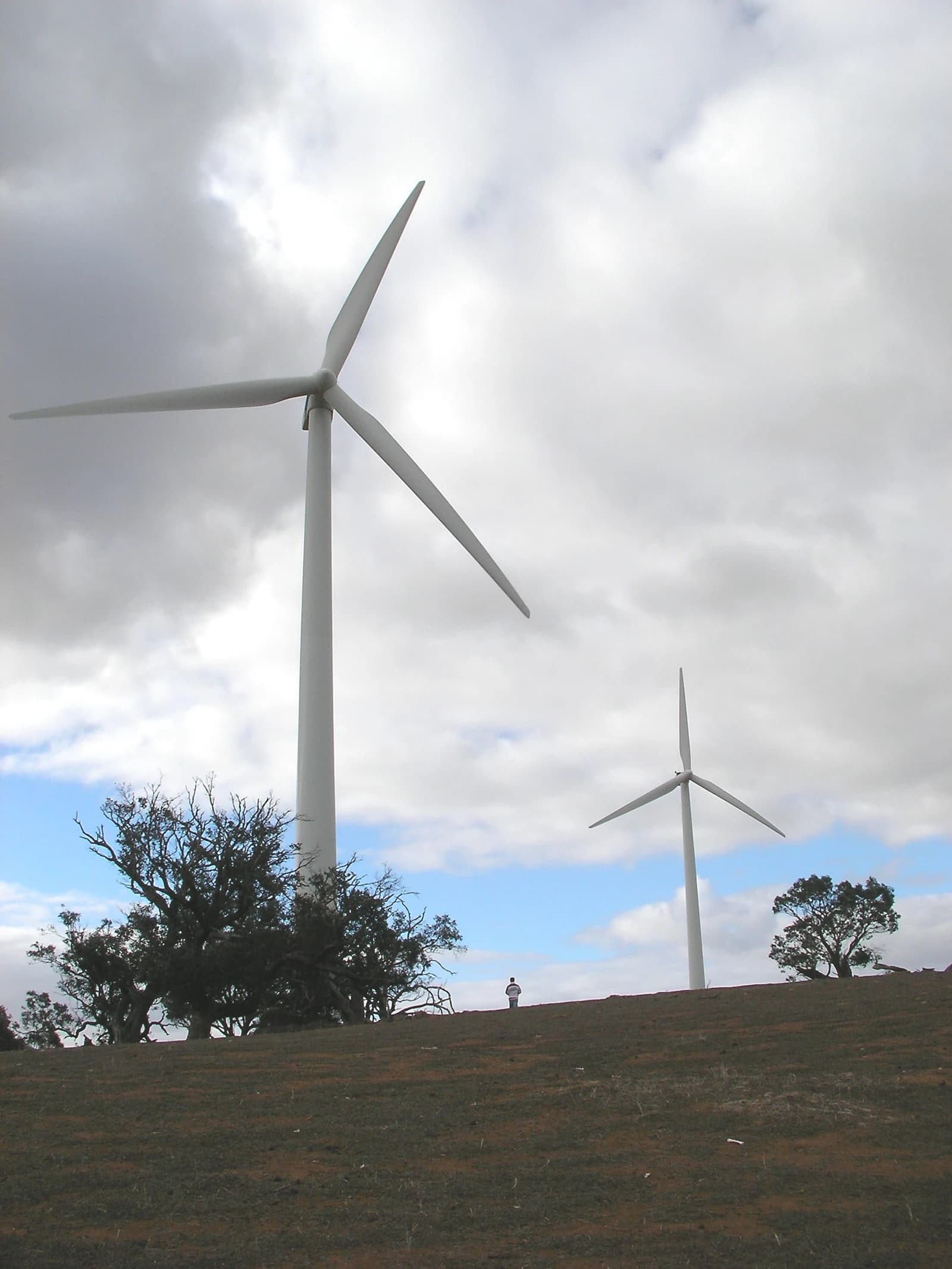 Two large wind turbines on a hill with a person standing between them.