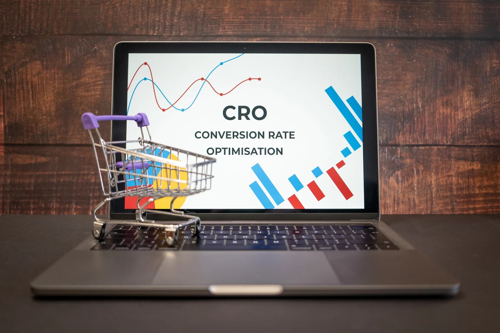 A miniature shopping cart rests on a laptop keyboard displaying charts and the words 'Conversion Rate Optimisation'.