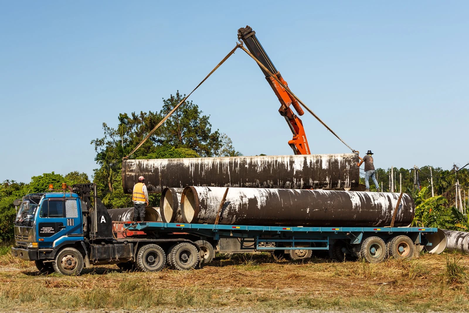 Construction workers loading large sections of pipe onto a flatbed truck using a hydraulic crane.