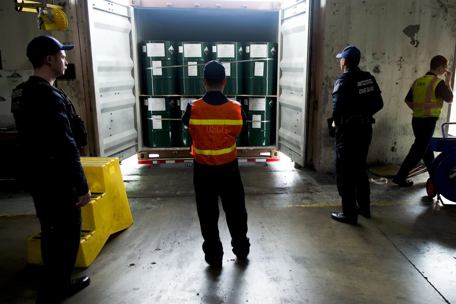 US Coast Guard personnel inspect green barrels inside an open shipping container at a port.