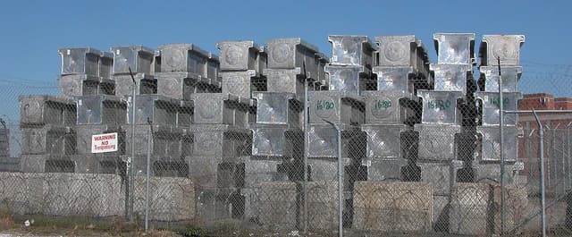 Stacked aluminium ingots in an outdoor storage area behind a fence.
