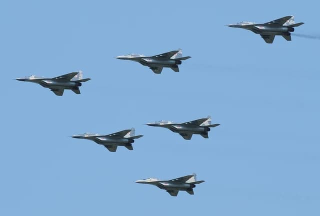 Six gray Mikoyan-Gurevich MiG-29A Fulcrum fighter jets flying in formation against a blue sky.