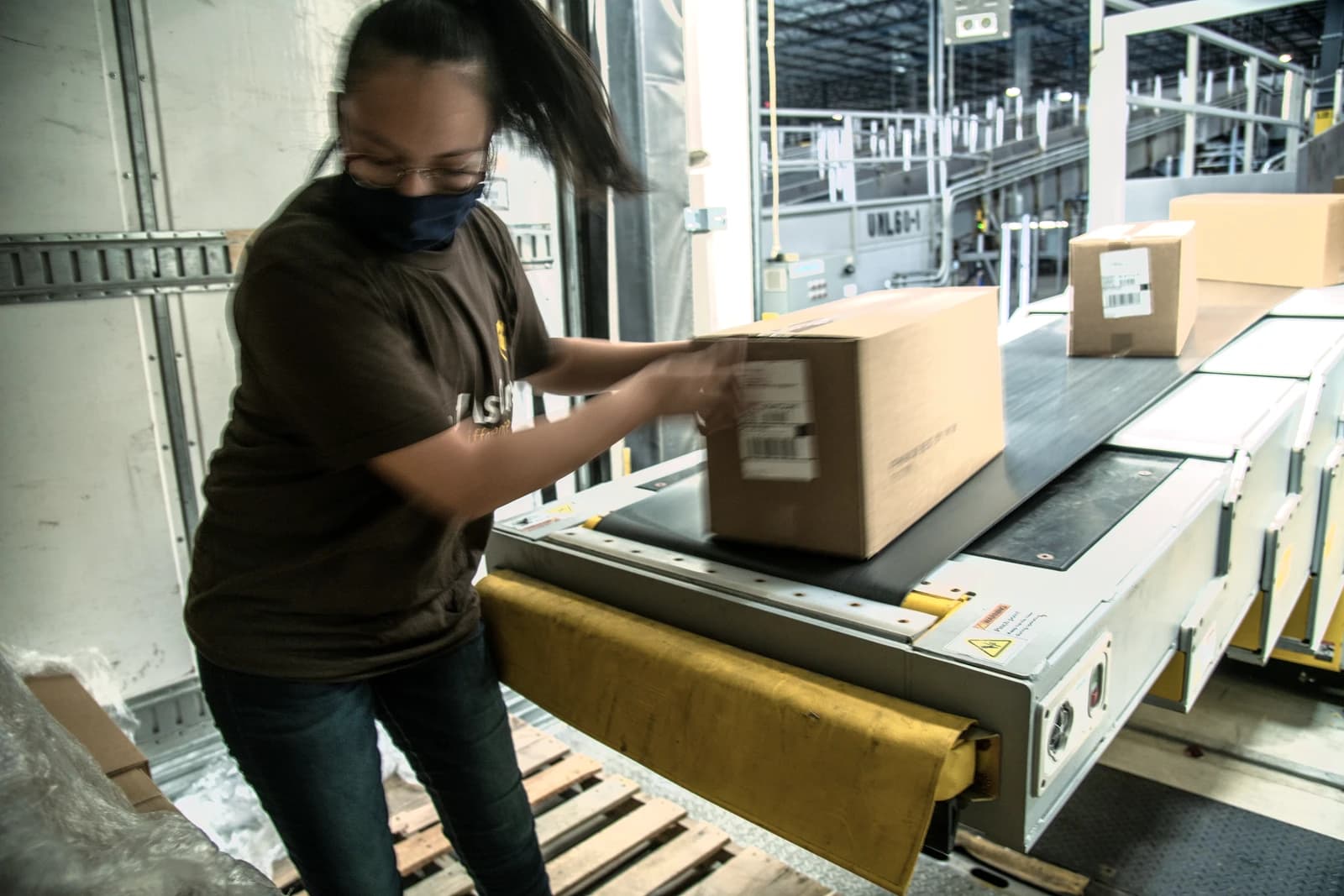 UPS worker, wearing face mask, unloads boxes from a semi-trailer truck onto a conveyor belt.
