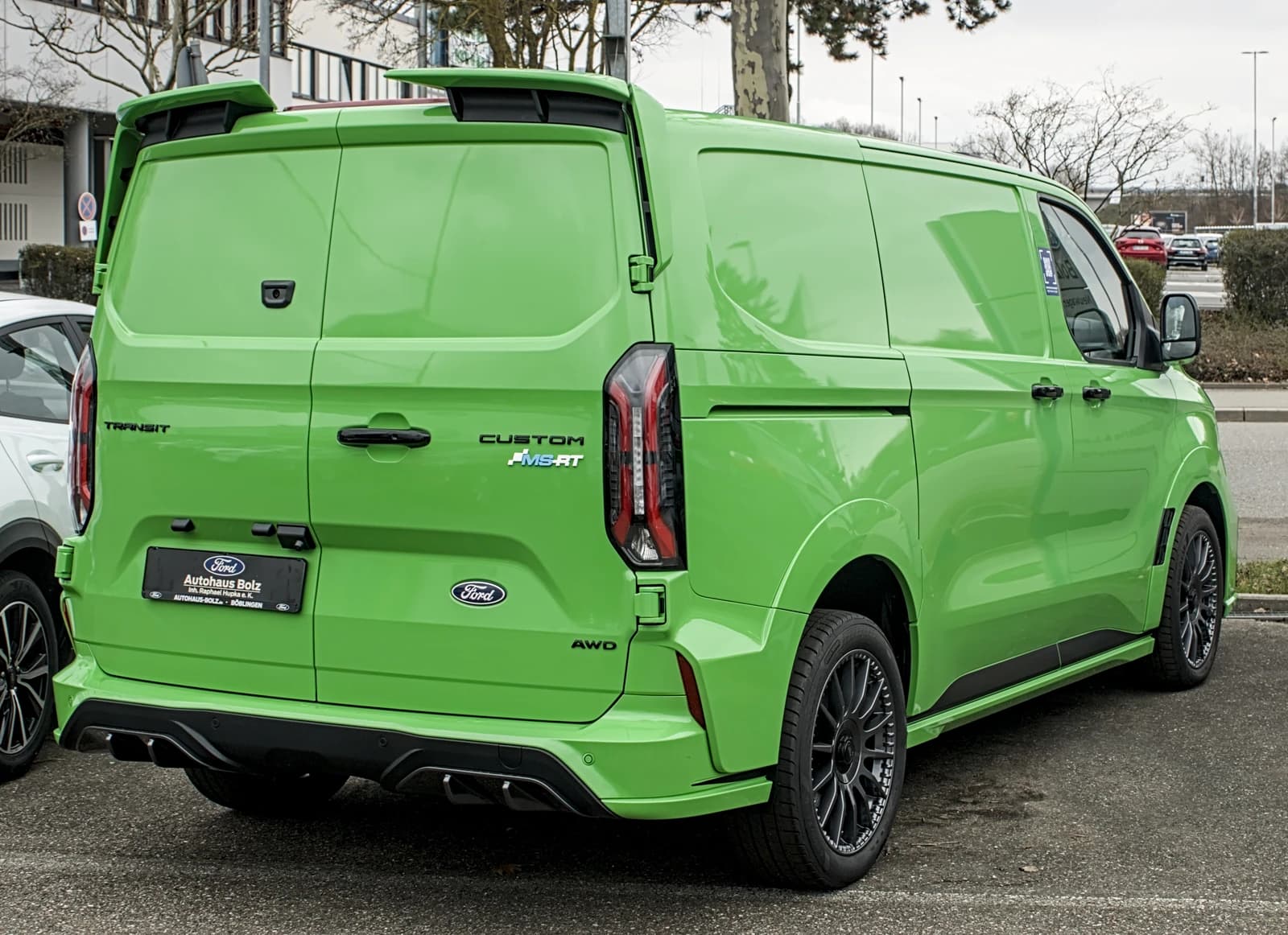 Green Ford E-Transit Custom MS-RT van parked in a lot, rear view.