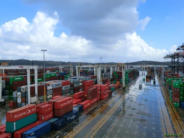 Shipping containers stacked at a port terminal under a cloudy sky.