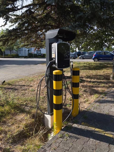 Electric vehicle charging station with a cable plugged into the unit, set against a background of parked cars.