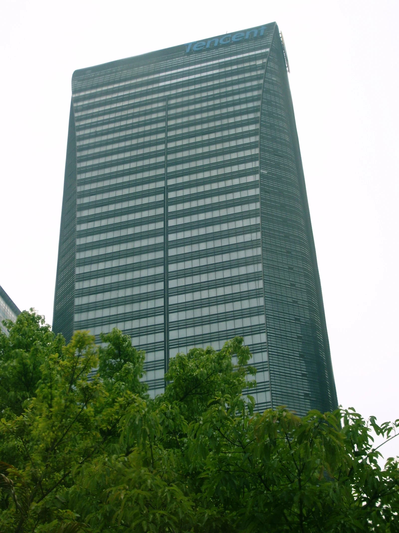 Tencent Headquarters, a tall, modern skyscraper, seen from behind green trees.