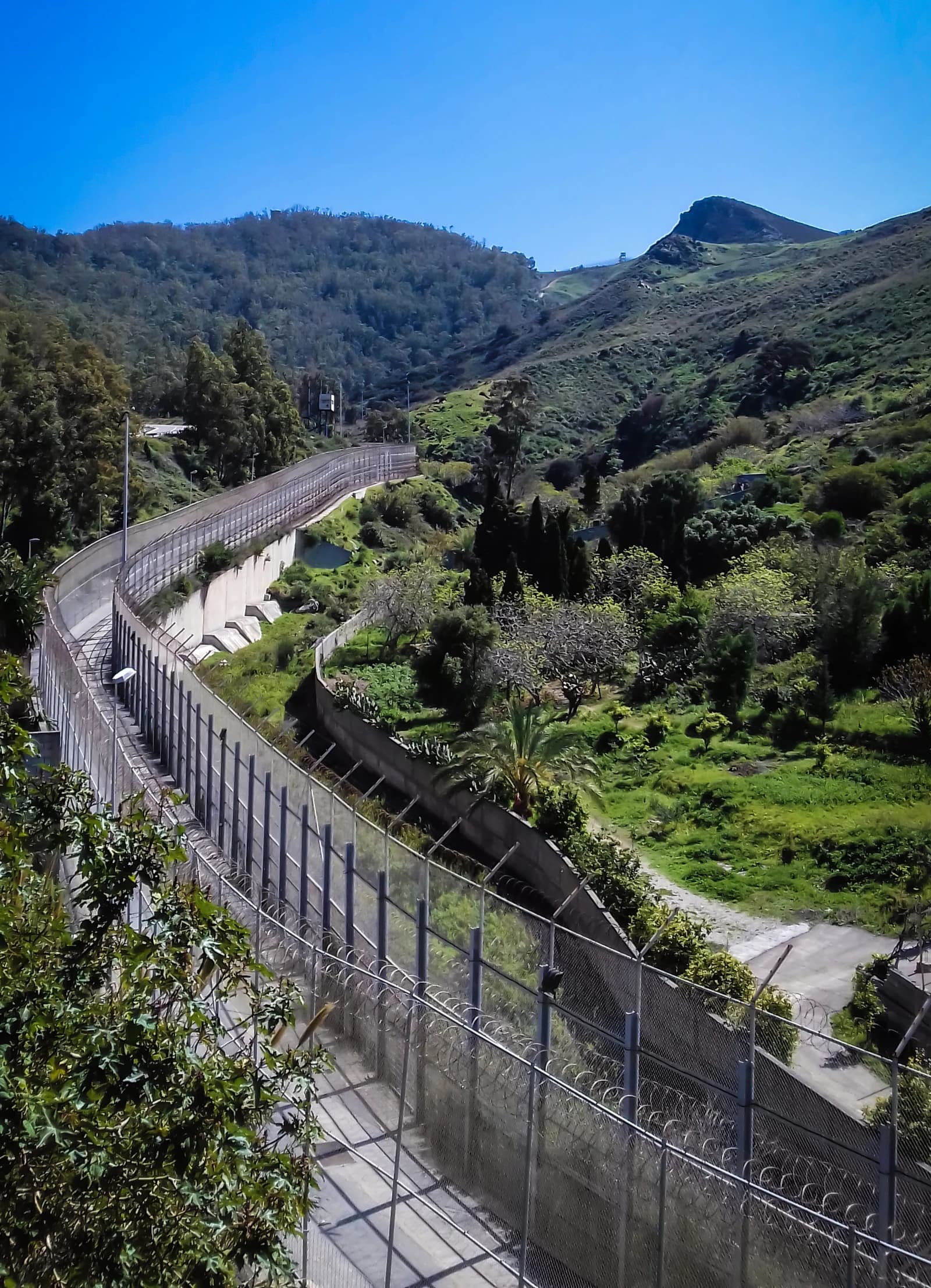 A long, winding border fence with barbed wire separates green hills under a blue sky.
