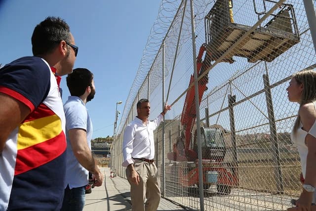 Javier Ortega, Spanish politician, points towards a high fence with barbed wire.
