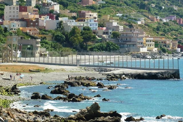A metal fence separating a beach and water from a hillside with buildings in Ceuta.