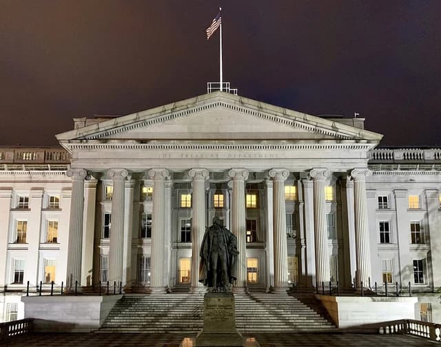 United States Treasury Building at night, illuminated, with a statue in front.