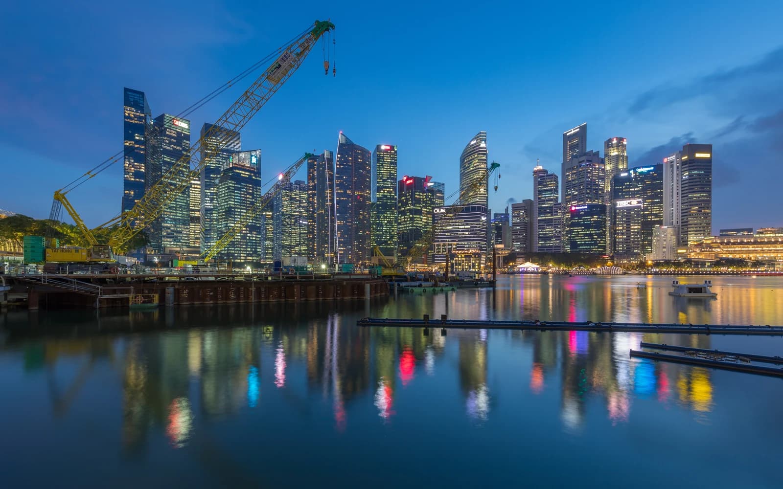 Singapore Central Business District skyline with illuminated buildings and construction cranes reflected in water.