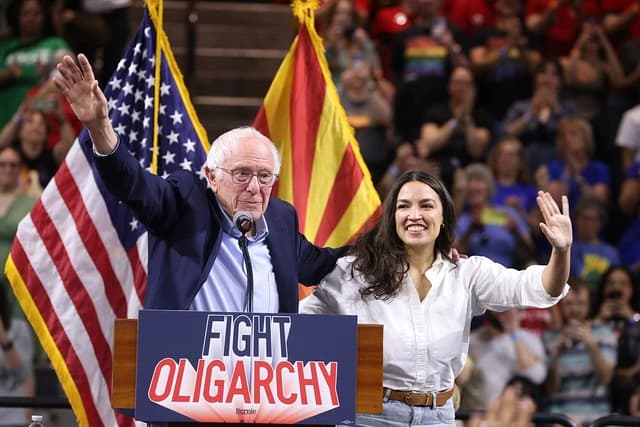 Bernie Sanders and Alexandria Ocasio-Cortez waving from a stage at an event.
