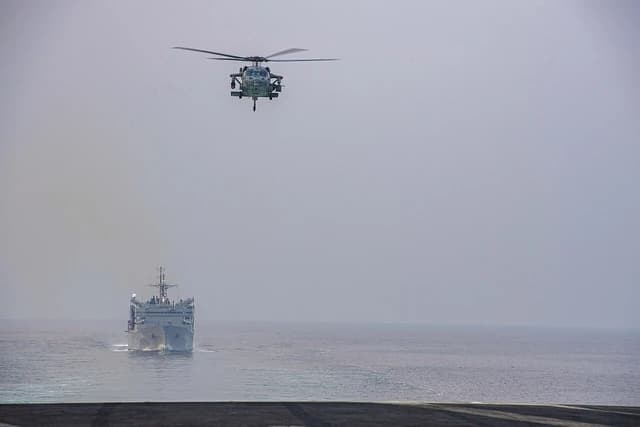 A US Navy helicopter flies high above a US Navy vessel navigating open water.