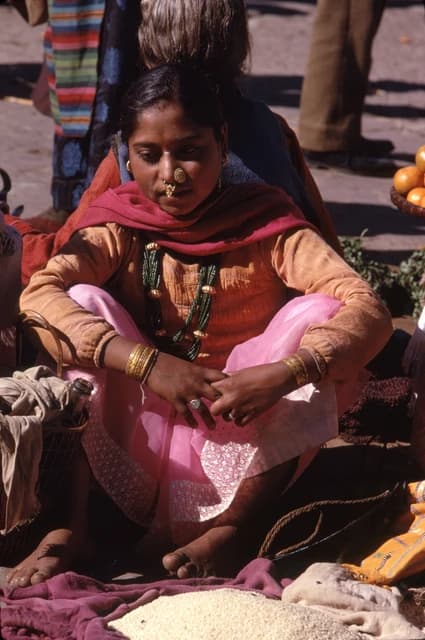 Woman wearing traditional clothing and jewelry, sitting next to a pile of rice.