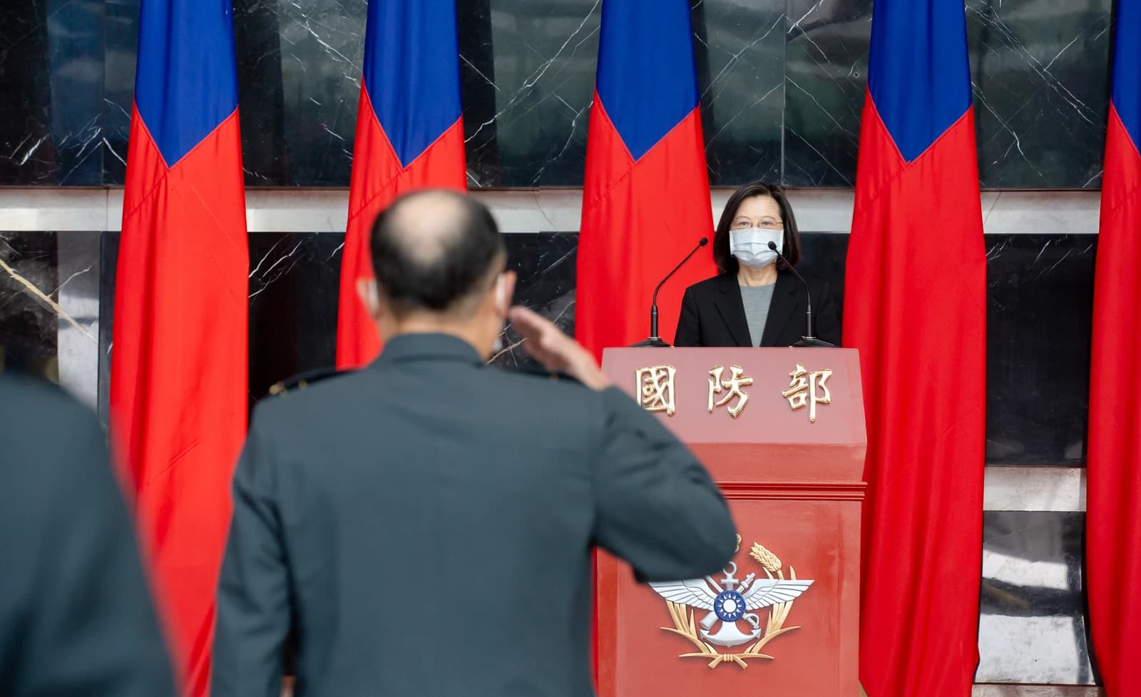 Taiwanese President Tsai Ing-wen speaking at a podium with an officer saluting her.