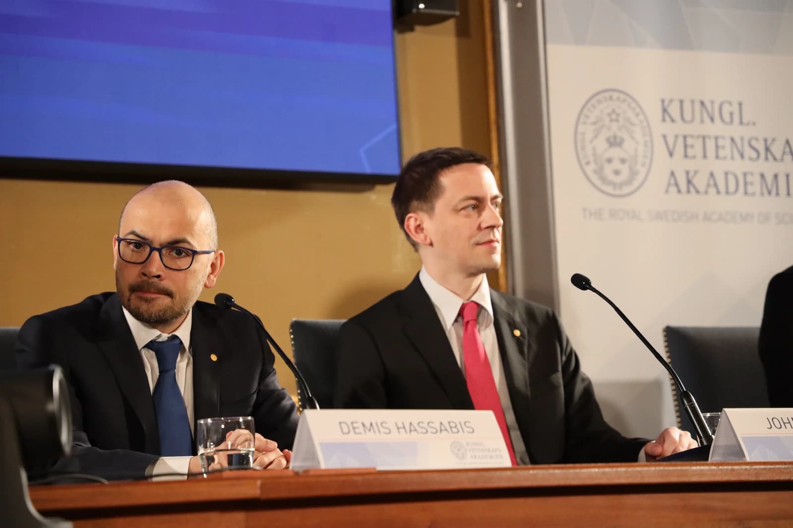 Demis Hassabis sitting at a table with a microphone and nameplate, looking towards the camera.