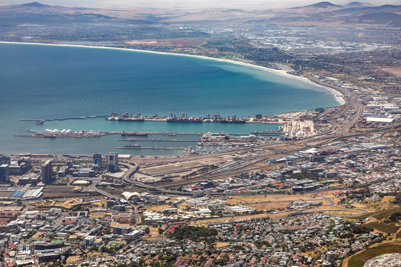 Aerial view of Cape Town city, harbor, and coastline under a bright sky.