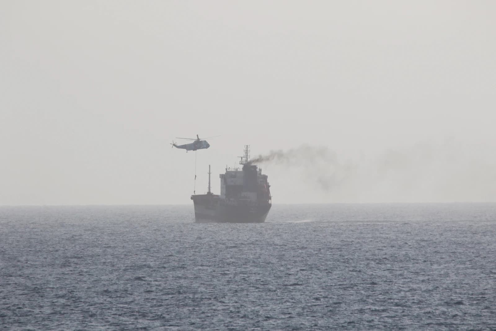 A helicopter hovering above a ship with smoke coming from its funnel on the open sea.
