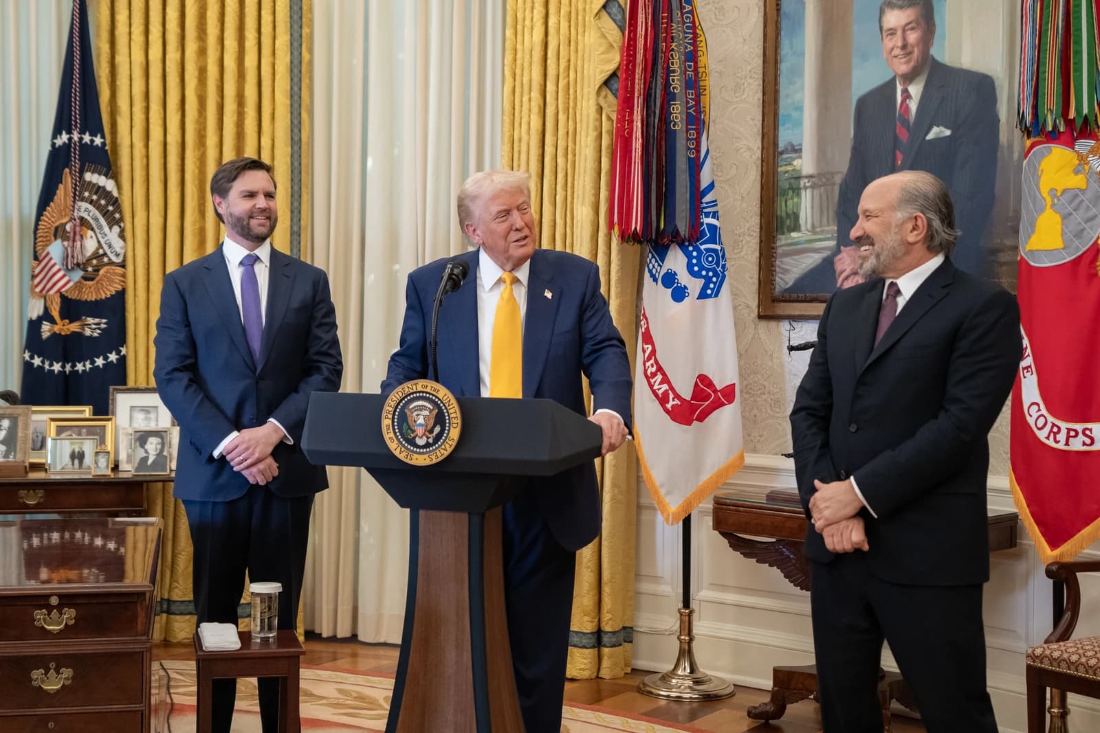 Donald Trump at a podium with JD Vance and Howard Lutnick standing nearby.