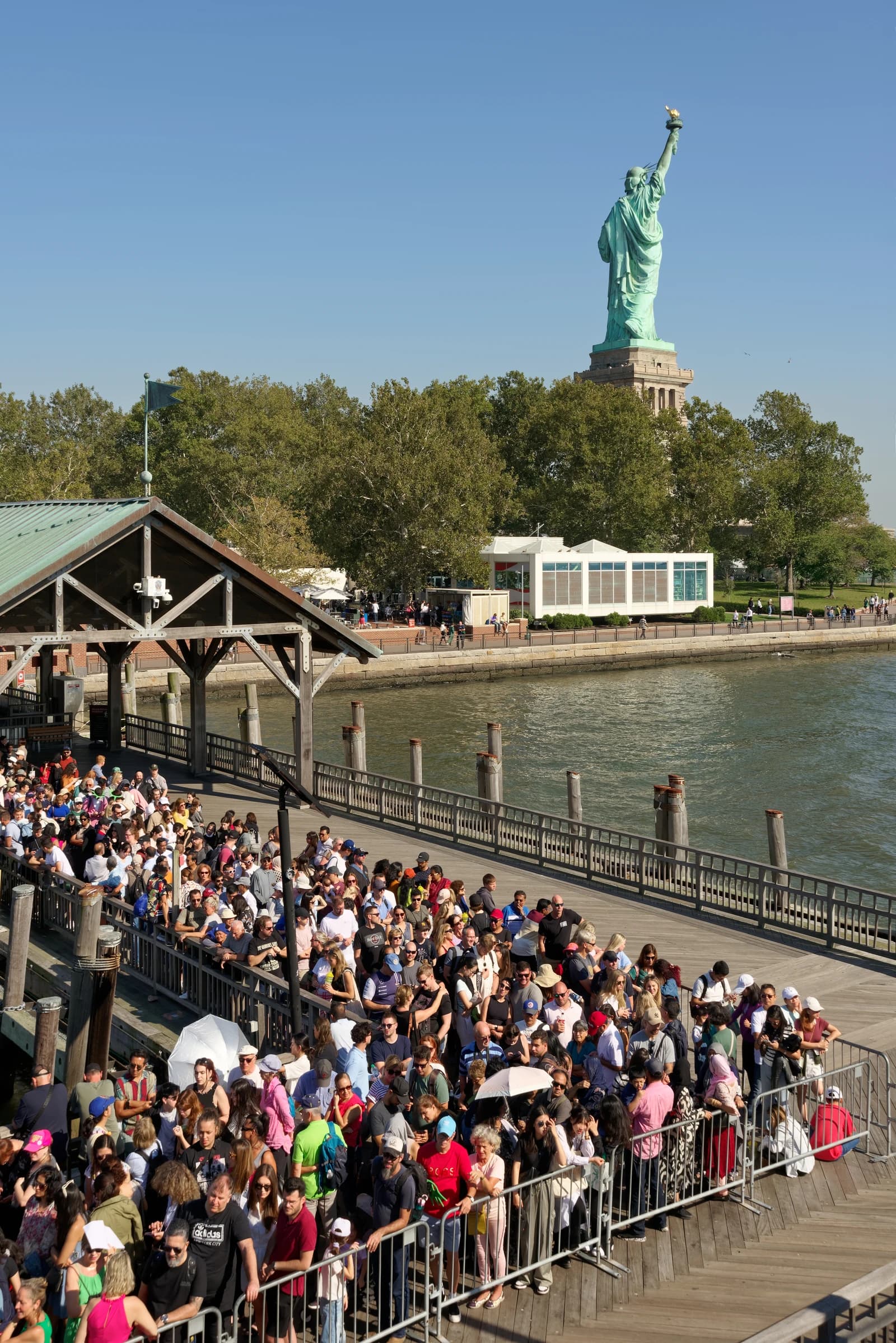 People wait in line at Liberty Island Ferry Terminal with the Statue of Liberty visible in the distance.