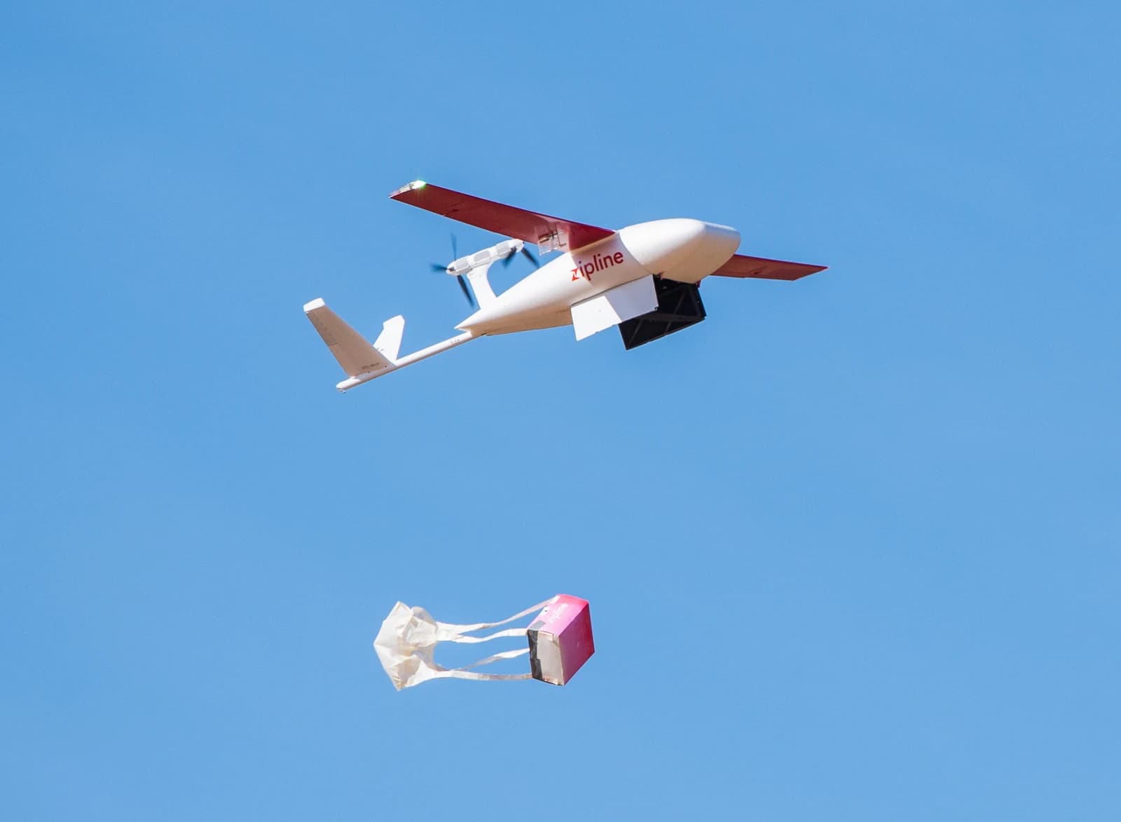 Zipline delivery drone with red wings and Zipline logo releasing a package from its underside.