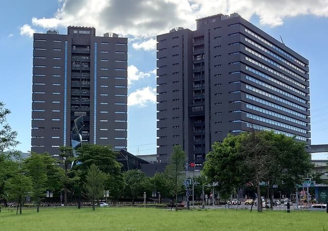 Xinzhuang Joint Office Tower, Executive Yuan, with two dark grey high-rise buildings and green trees.