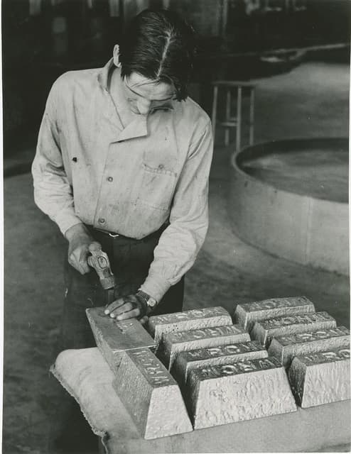 Man holding a hammer, stamping a gold ingot on a table with other ingots.